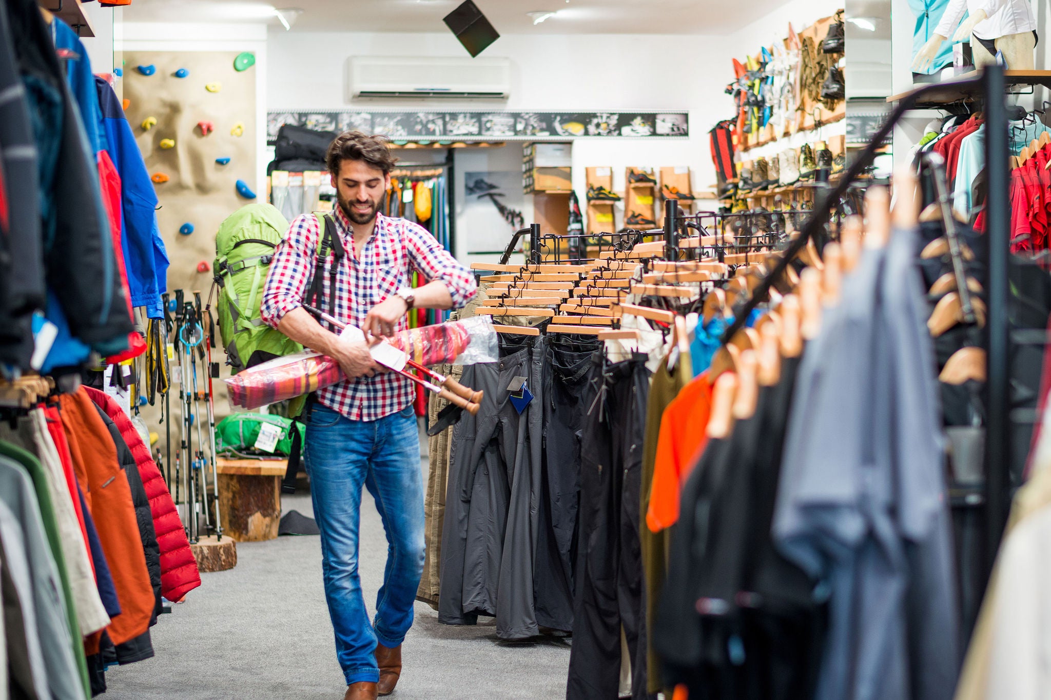 Bearded man in sports and outdoor equipment store looking to buy hiking equipment. He is holding green backpack, exercise mat, drinking bottle and hiking poles.
